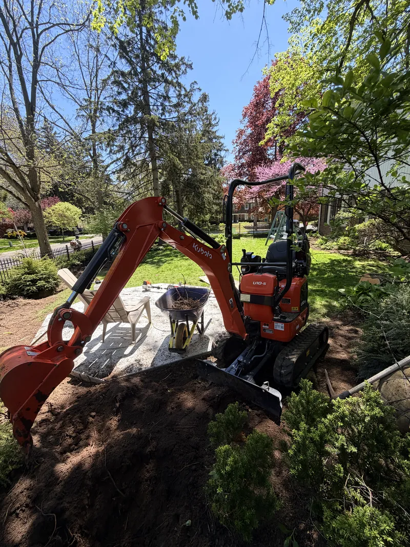 Operator working a compact excavator on a front garden excavation with a redbud in flower behind.
